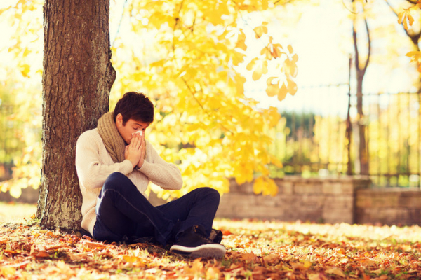 photo of a man sitting on the ground with his back against a tree holding a tissue to his face and blowing his nose; ground is covered in leaves indicating fall season